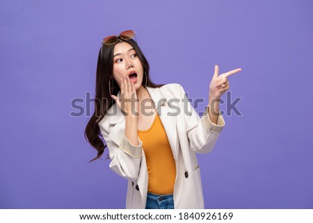 Waist up portrait of shocked pretty Asian woman pointing hands to empty space aside on isolated purple studio background