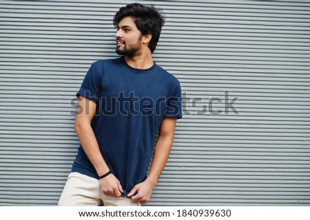 Stylish casual indian man wear blue t-shirt posing against grey wall.