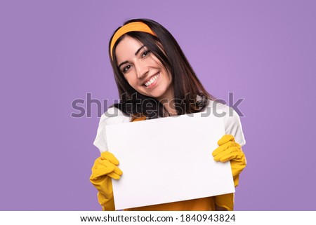 Positive young female in rubber gloves demonstrating blank white paper sheet while representing housekeeping and housework concept against purple background