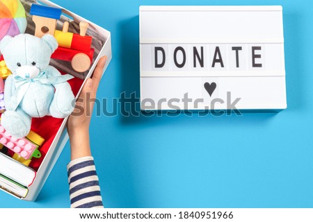 Hands holding donation box with toys books clothing and lightbox with text donate on light blue background. Top view