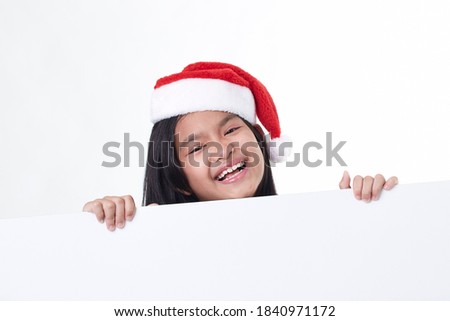 Portrait of a happy little girl wearing Santa hat posing behind a white panel isolated on white background holding a blank board Christmas billboard