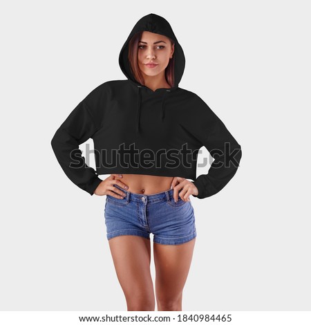 Template female crop top on a beautiful girl in a hood with hands on a belt, an empty black hoodie for design presentation. Mockup of casual fashion clothes on a white background in the studio.