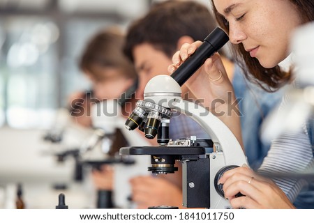 Close up of young woman seeing through microscope in science laboratory with other students. Focused college student using microscope in the chemistry lab during biology lesson. Royalty-Free Stock Photo #1841011750