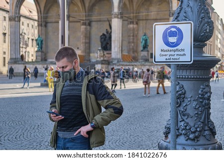 Man in a protective face mask near the sign that warns people to cover their mouth and nose at Odeon square in Munich, Germany. Man wearing a mask is holding his phone at Odeonplatz Royalty-Free Stock Photo #1841022676