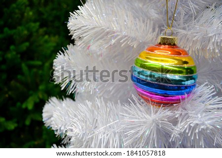 Bright glass rainbow colored Christmas ball, bauble hanging on a white artificial christmas tree. On background of green christmas tree. The concept of holiday, Christmas, symbol of the rainbow