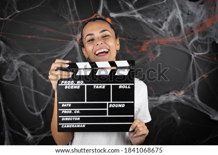 Young beautiful woman over black background with cobwebs and spiders holding clapperboard very happy having fun