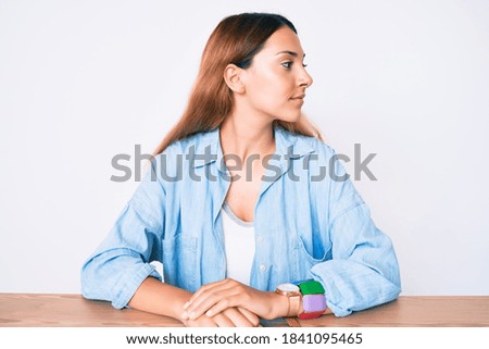 Young brunette woman sitting on the table wearing casual clothes looking to side, relax profile pose with natural face with confident smile. 