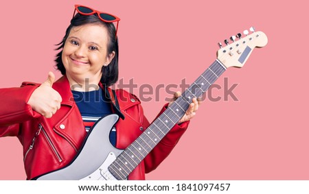 Brunette woman with down syndrome playing electric guitar smiling happy and positive, thumb up doing excellent and approval sign 
