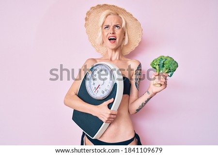 Young blonde woman with tattoo wearing bikini holding weighing machine and broccoli angry and mad screaming frustrated and furious, shouting with anger looking up. 