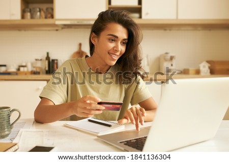 Cute Arabic girl transferring money with online banking using wireless internet connection on laptop. Stylish young woman holding debit card entering personal payment data, purchasing goods distantly