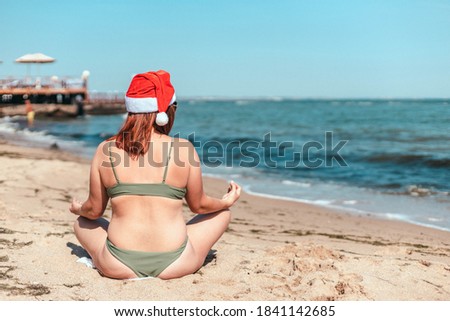 Woman in Santa Claus hat sitting on sand of the beach. Woman in swimsuit sitting in lotus position and enjoying the beautiful view