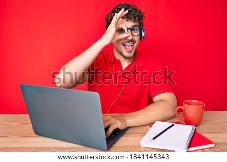 Young caucasian man with curly hair working at the office drinking a cup of coffee smiling happy doing ok sign with hand on eye looking through fingers 