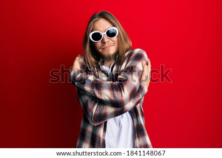 Handsome caucasian man with long hair wearing hipster shirt wearing sunglasses hugging oneself happy and positive, smiling confident. self love and self care 