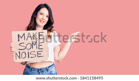 Beautiful young brunette woman holding make some noise banner smiling happy pointing with hand and finger to the side 