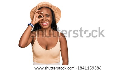 Young black woman wearing summer hat doing ok gesture with hand smiling, eye looking through fingers with happy face. 