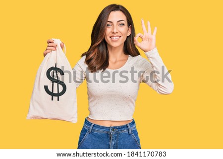 Young brunette woman holding dollars bag doing ok sign with fingers, smiling friendly gesturing excellent symbol 
