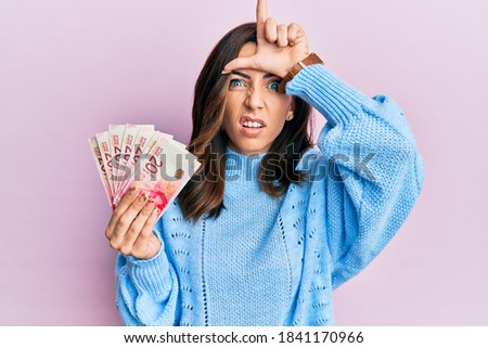 Young brunette woman holding 20 israel shekels banknotes making fun of people with fingers on forehead doing loser gesture mocking and insulting. 
