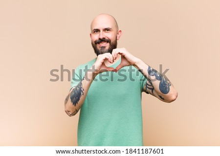 young bald and bearded man smiling and feeling happy, cute, romantic and in love, making heart shape with both hands