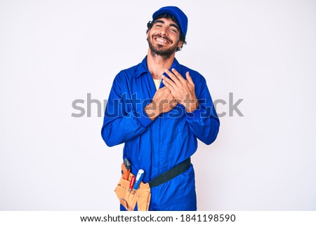 Handsome young man with curly hair and bear weaing handyman uniform smiling with hands on chest with closed eyes and grateful gesture on face. health concept. 