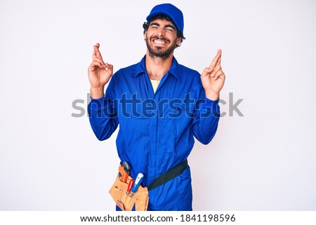 Handsome young man with curly hair and bear weaing handyman uniform gesturing finger crossed smiling with hope and eyes closed. luck and superstitious concept. 