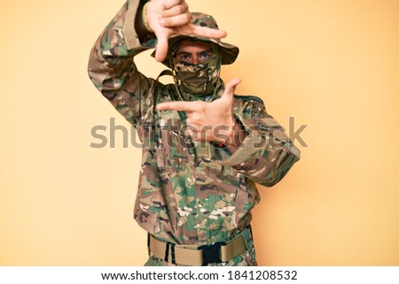 Young handsome man wearing camouflage army uniform and balaclava smiling making frame with hands and fingers with happy face. creativity and photography concept. 