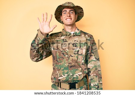 Young handsome man wearing camouflage army uniform showing and pointing up with fingers number five while smiling confident and happy. 