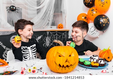 Happy Halloween! Attractive young boy with his brother are preparing for Halloween party. Brothers in costumes are having fun with pumpkins and cupcakes.