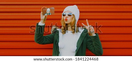 young woman taking selfie picture by phone wearing a knitted hat, sweater on red background