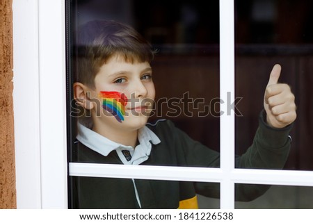 Lonely school kid boy sitting by window with rainbow with colorful colors on face. Child during pandemic coronavirus quarantine. Children make and paint rainbows around the world