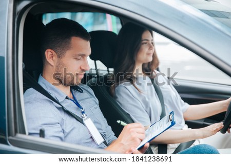Learning to drive a car. Male instructor and female student practicing in driving. Young man notice results of test in his paper. Cheerful woman satisfied with driving lessons. Royalty-Free Stock Photo #1841238691