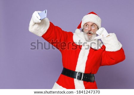 Shocked Santa Claus man in Christmas hat red coat glasses doing selfie shot on mobile phone pointing thumb on himself isolated on violet background. Happy New Year celebration merry holiday concept