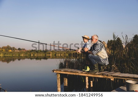 Senior man sitting on jetty fishing in lake with grandson