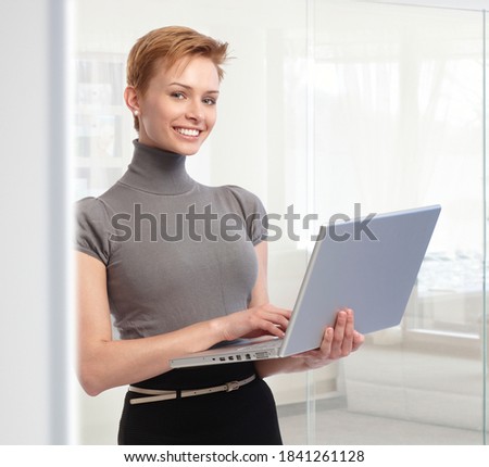 Young attractive elegant caucasian businesswoman standing with laptop computer at office. Working, looking at camera, toothy smile.  .