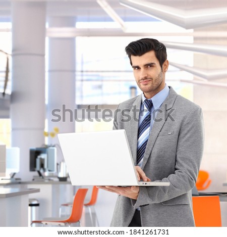 Confident caucasian businessman working on laptop computer at bright business office. Suit, small smile, standing.