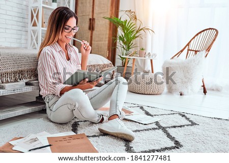 Serious pretty woman with dark hair, writes in notebook list to do in morning, has many plans for coming weekends, poses on floor indoor. Copywriter works at home Royalty-Free Stock Photo #1841277481