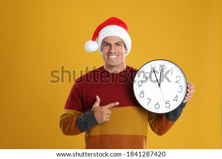 Man in Santa hat with clock on yellow background. New Year countdown