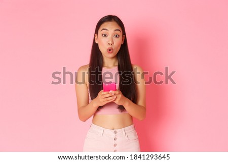 Technology, communication and online lifestyle concept. Intrigued asian trendy girl looking amused with special offer in internet store, holding mobile phone and pouting curious, pink background