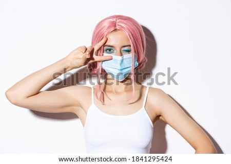 Coronavirus, social distancing and lifestyle concept. Close-up of beautiful cheerful girl in medical mask and pink wig, staying positive, showing peace sign and winking joyfully