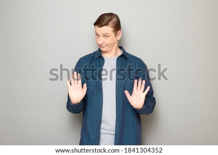 Studio portrait of funny skeptical mature man wearing blue shirt, showing palms at camera, rejecting something, saying no or enough, or asking not to worry and panic, standing over gray background