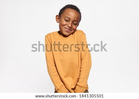 Horizontal shot of cute handsome dark skinned little boy shrugging shoulders feeling embarrassed with uncomfortable question, looking down with shy timid smile. Human emotions and body language Royalty-Free Stock Photo #1841305501