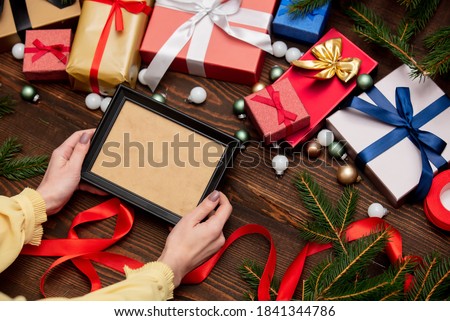 female hand holds picture frame near with christmas gifts and pine branches with baubles on wooden table