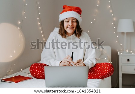 Young teen girl in red santa hat shopping online buying Christmas gifts paying by credit card using laptop sitting on bed in her bedroom. Child internet security 