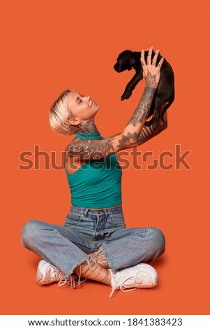 Cheerful young adult woman wearing in casual wear, holding cute puppy in hands, smiling wide and sitting on floor in studio at orange background with copy space