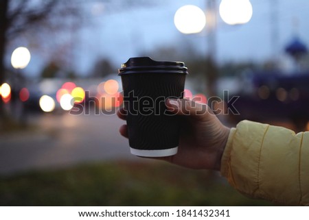 Holding a cup of coffee and raise my hand to take a picture with a blurry background. Black cup of coffee without logo in hand.