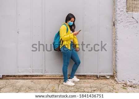 happy girl wearing yellow jacket using her smartphone in the street with face mask.