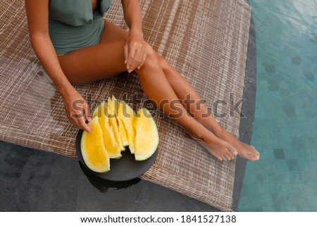 Girl eat slices of yellow watermelon in the blue pool, slim legs. Tropical fruit diet. Summer holiday idyllic island