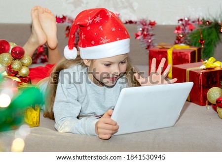 Christmas greetings online. A smiling blonde girl uses her mobile tablet for greeting and waves her hand. Greeting video calls to friends, relatives, and parents