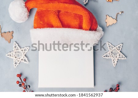 White square canvas board in Santa hat on grey background with Christmas decoration. Mockup poster frame for Christmas message or design.