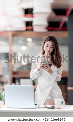 Portrait of young asian woman using smartphone while standing at her office desk in modern office.