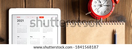 a tablet with an open calendar for 2021 year, a red alarm clock, and a craft paper notebook on a wooden boards table background. banner Royalty-Free Stock Photo #1841568187